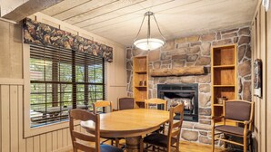 A cozy dining area with a stone fireplace, wooden chairs, and a large window with blinds. - 🏡 2BR Branson Cabin Retreat | Pools, Hot Tub, Pet-Friendly (Branson)