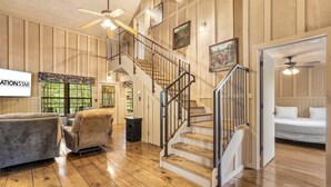 A spacious living area with a wooden floor, a staircase with a metal railing, and a bed in the adjacent room. - 🏡 2BR Branson Cabin Retreat | Pools, Hot Tub, Pet-Friendly (Branson)