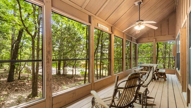 A covered outdoor seating area with wooden furniture and large windows overlooking a forest. - 🏡 2BR Branson Cabin Retreat | Pools, Hot Tub, Pet-Friendly (Branson)