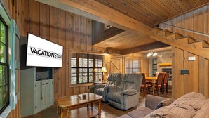 Een houten interieur met een televisie waarop 'VACATIONSTAR' staat, een bank, een relaxfauteuil, een eettafel met stoelen en een trap.