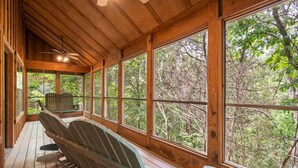 Een houten terras met Adirondack-stoelen en grote ramen met uitzicht op een bos.