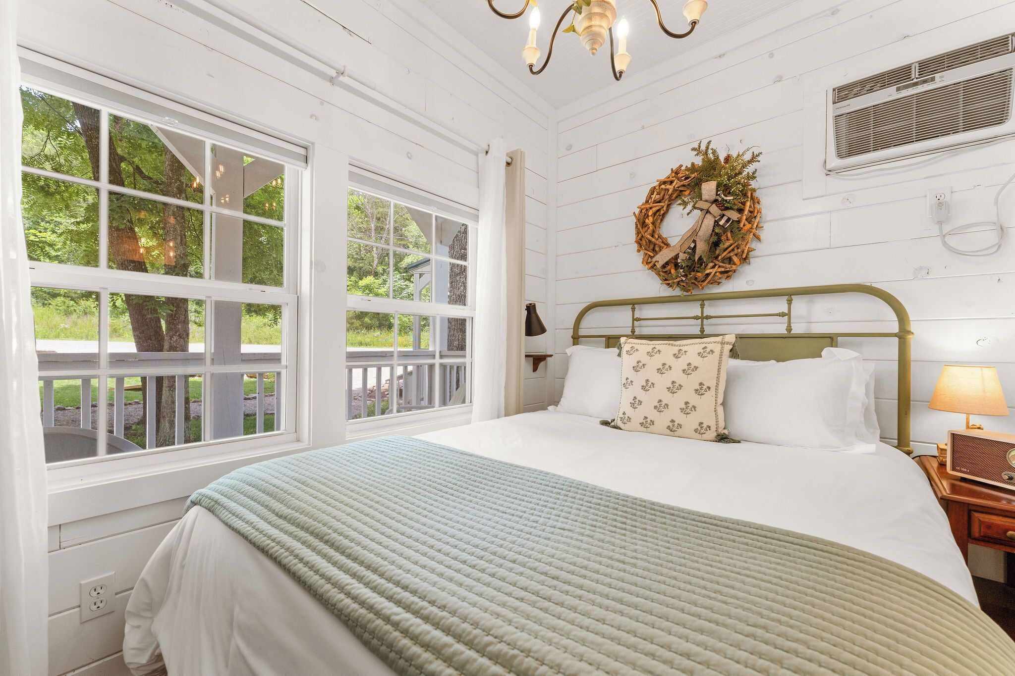 A bedroom with a large window, a bed with a green blanket, a nightstand with a lamp, and a wreath on the wall.