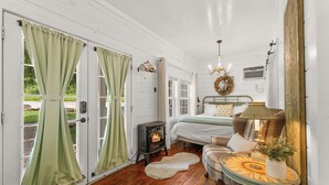 A cozy bedroom with a wood-burning stove, a bed with a green and white duvet, a small table with a sunflower design, and a door with green curtains.