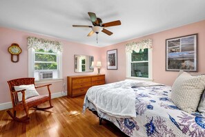 A bedroom with a wooden bed, a rocking chair, a ceiling fan, and a window with a view of trees. - Family & Pet-Friendly Cape Escape Near Beach (Hyannis)
