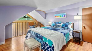 A bedroom with a sloped ceiling, a bed with a blue and white beach-themed bedspread, a wooden bench, and a small table with a lamp. - Family & Pet-Friendly Cape Escape Near Beach (Hyannis)