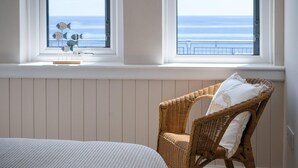 Ein Schlafzimmer mit einem Bett, einem Korbstuhl mit Kissen und Blick auf das Meer durch zwei Fenster.