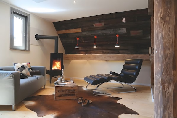 A cozy living room with a sofa, a leather chair, a fireplace, and a wooden ceiling.