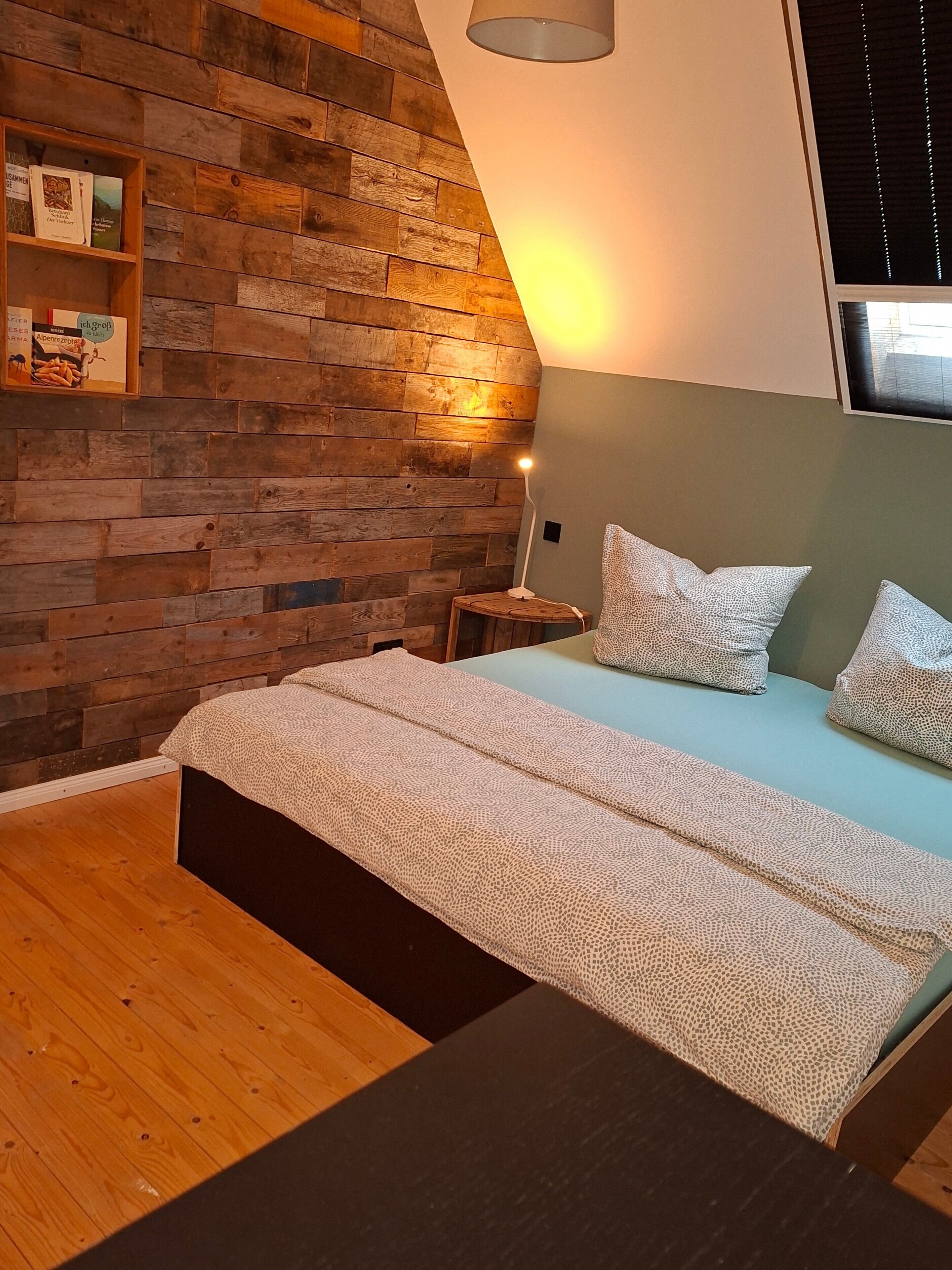 A bedroom with a sloped ceiling, a wooden wall, a bedside table with a lamp, and a bed with patterned pillows.