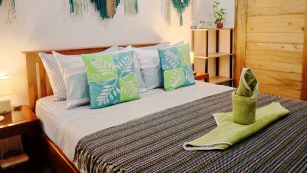 A neatly made bed with patterned pillows, a woven blanket, and a green towel. There are macramé wall hangings above the bed and a wooden door to the right. - Amwani Residence - Kalikasan Homes (Puerto Princesa)
