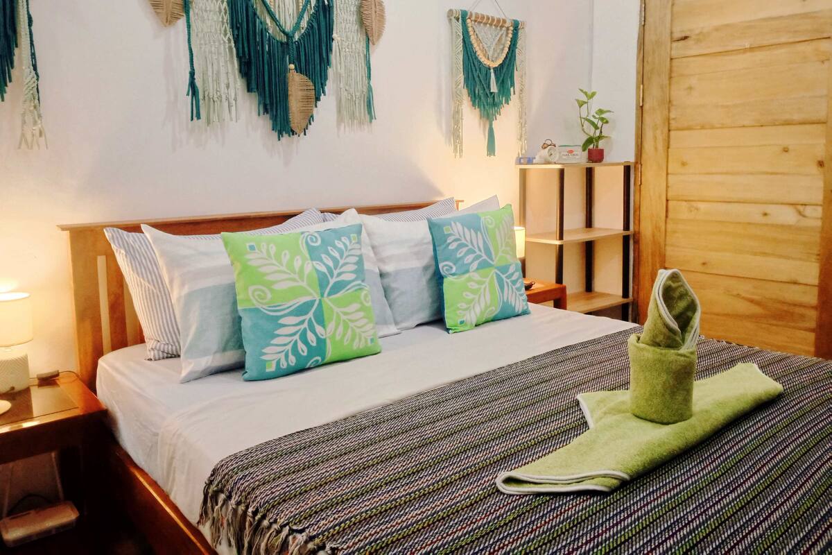A neatly made bed with patterned pillows, a woven blanket, and a green towel. There are macramé wall hangings above the bed and a wooden door to the right.