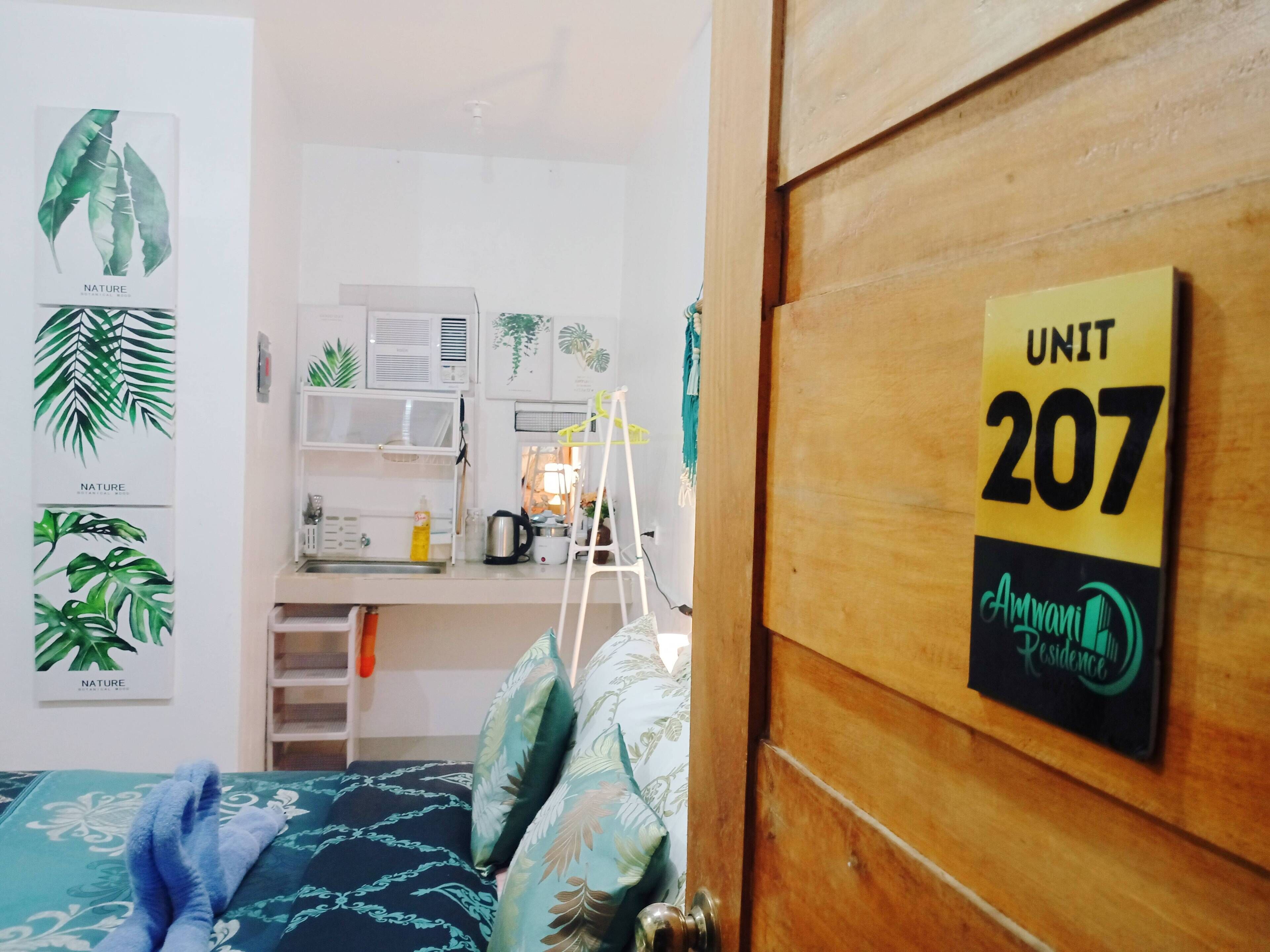 Ein Schlafzimmer mit einem Bett, einer Holztür mit Hinweisschild für die Zimmernummer, einem Schreibtisch mit Computer, Regalen und einem Fenster.