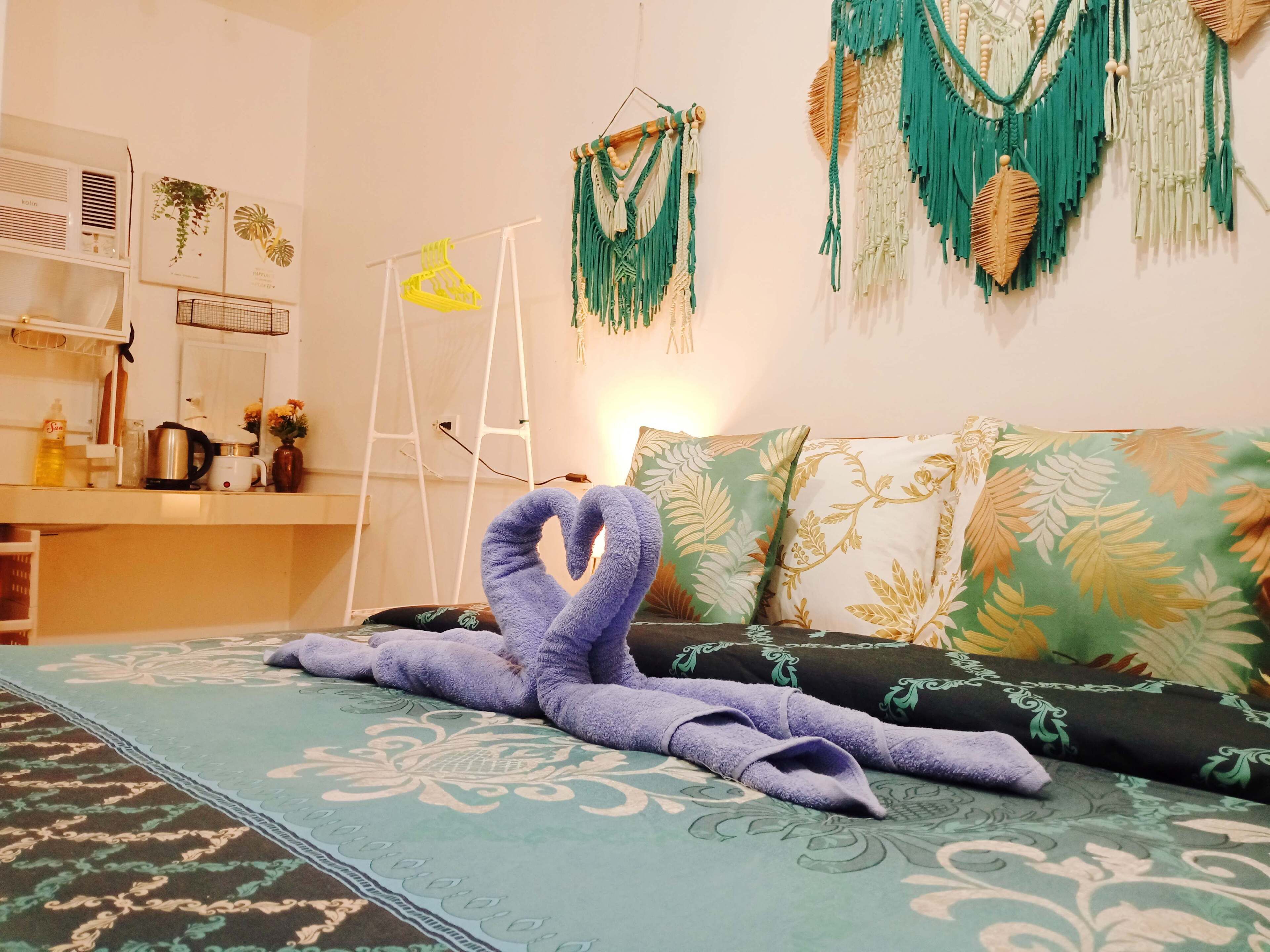 A neatly made bed with a purple towel, a green and gold patterned sofa, and macramé wall hangings.