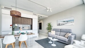 Una sala de estar moderna con un sofá gris, un comedor con sillas blancas y una cocina con armarios blancos.