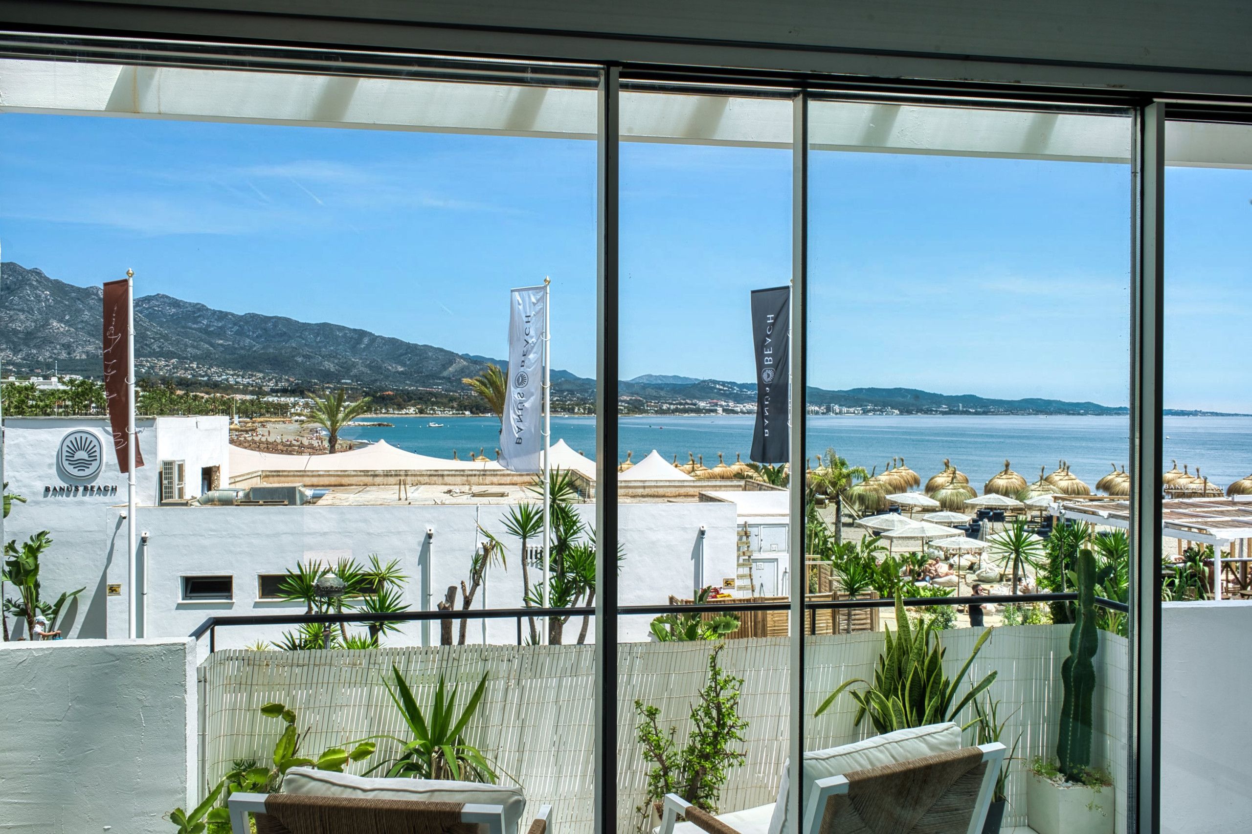 Un balcon avec vue sur une plage et des montagnes. On peut voir des drapeaux et des parasols sur la plage.