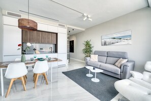 Una sala de estar moderna con un sofá gris, un comedor con sillas blancas y una cocina con armarios blancos.