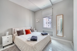 A modern bedroom with a large bed, a bedside table, a nightstand, a window with bars, and a mirror on the wall.