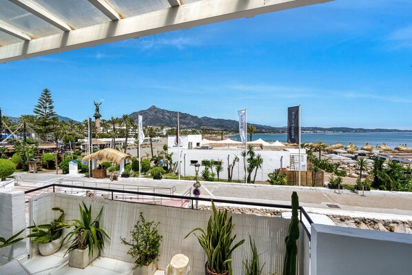 A coastal hotel with a terrace overlooking the beach and sea, featuring potted plants and a clear view of the mountains in the background.