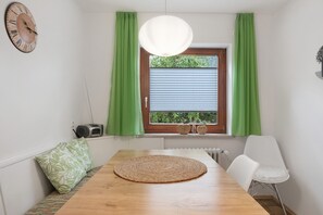 A dining area with a wooden table, a woven placemat, a white chair, a clock, and a window with green curtains. - Apartment '1' with Mountain View, Private Terrace and Wi-Fi (Sankt Peter)