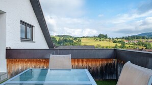 A balcony with a glass table and chairs, overlooking a green landscape with houses and hills.