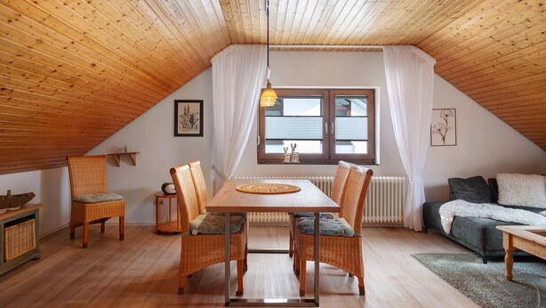 A cozy attic room with a wooden ceiling, a dining table with chairs, a sofa, and a window with curtains.