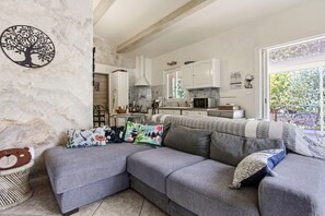 Un salon moderne avec un canapé d’angle gris, une décoration murale représentant un arbre, un fauteuil en osier pour chat et une cuisine en arrière-plan.