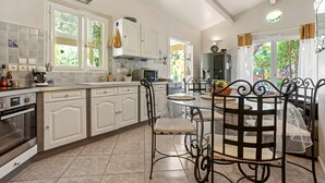 A kitchen with white cabinets, a dining table with chairs, and a view of greenery outside. - Villa 'Palmiers En Couleurs' with Mountain View, Private Pool, and Wi-Fi (Cuers)