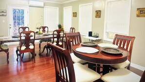 Ein Esszimmer mit einem runden Holztisch, sechs Stühlen und einem Tisch mit Glasplatte. Weiße Jalousien, ein kleiner weißer Beistelltisch und eine Tür mit bleiverglastem Fenster sind ebenfalls vorhanden.
