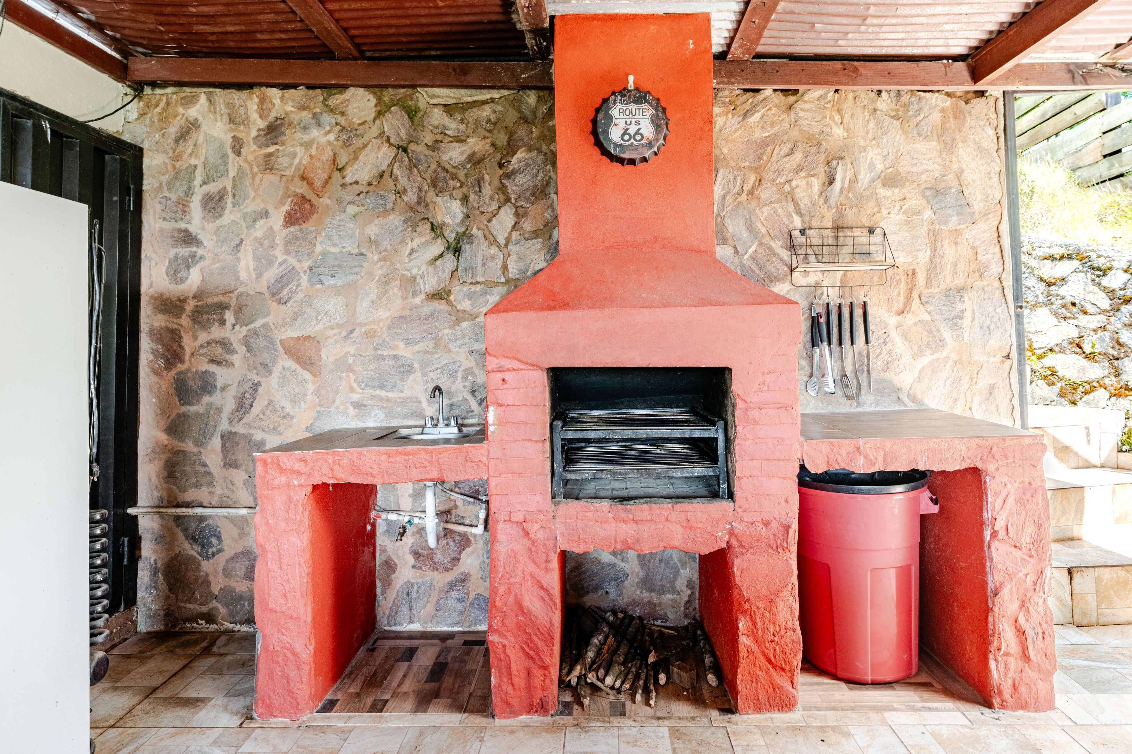 Una pared de piedra exterior con una parrilla de ladrillo rojo y un soporte de metal para utensilios de barbacoa.