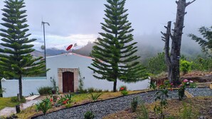 Un edificio bianco con una porta in legno, circondato da un giardino con piante e fiori diversi, e un vialetto sterrato che vi conduce.