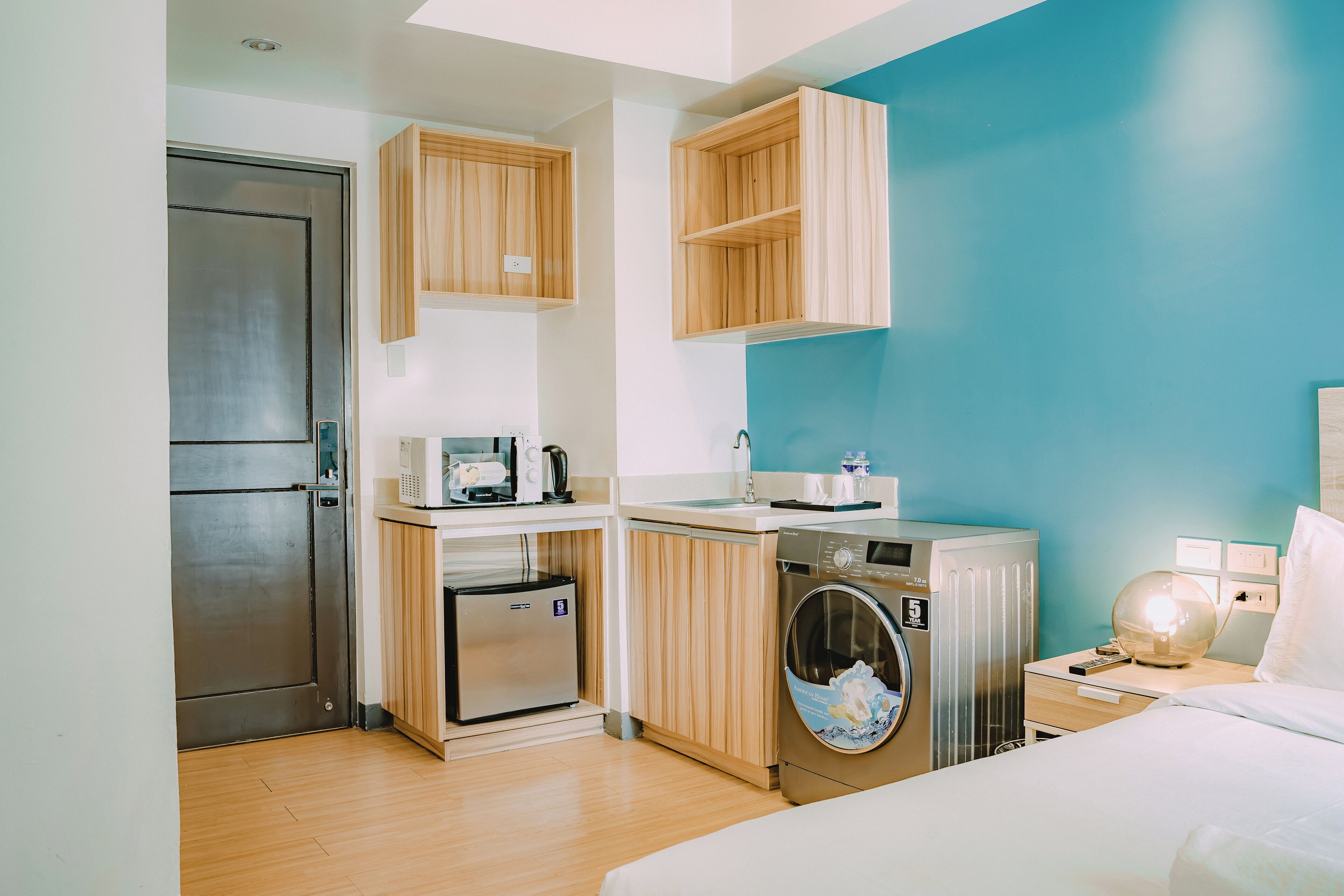 A compact hotel room with a kitchenette, a washing machine, a bed, and a nightstand with a lamp.