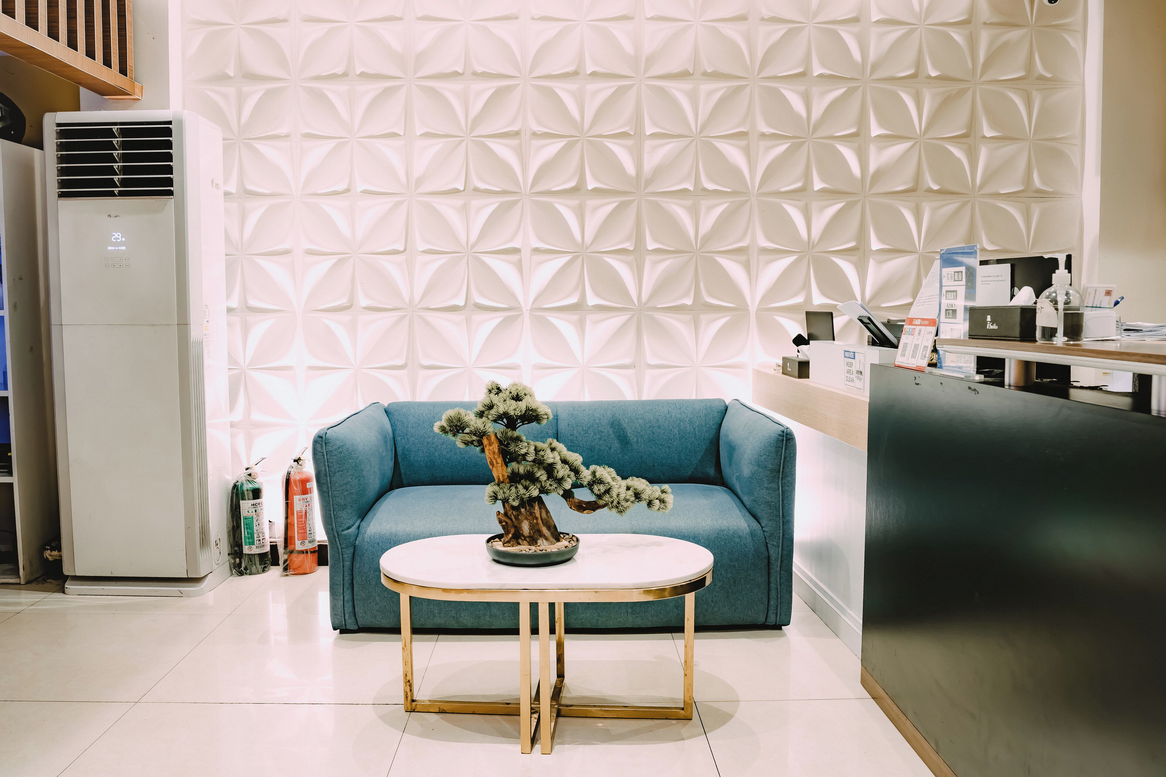 Un espace d’accueil avec un canapé bleu, une table ronde ornée d’un bonsaï, un mur blanc à texture et un comptoir de réception sur lequel sont disposés divers objets.