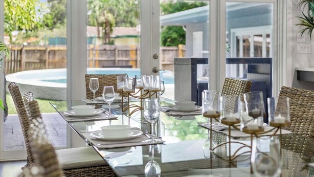 A dining area with a glass table, wicker chairs, and a view of a pool and garden. - Your Hollywood Vacation Home (Hollywood)