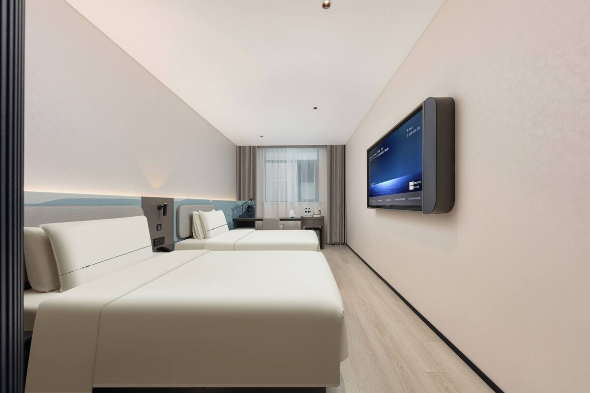 A modern hotel room with two beds, a desk, a flat-screen TV mounted on the wall, and a large window with curtains.