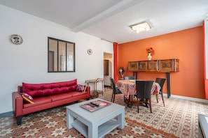 Uma sala de estar com um sofá vermelho, uma mesa de centro branca, uma área de jantar com mesa e cadeiras, e um piso estampado.