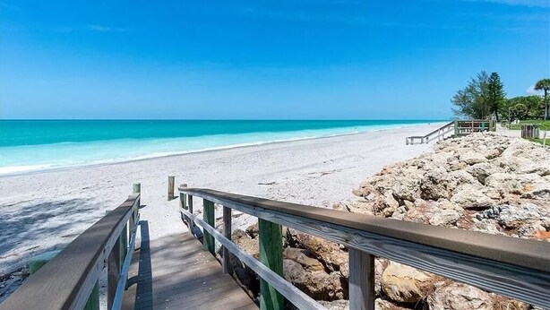 Un paseo marítimo de madera que conduce a una playa con agua turquesa cristalina y una costa rocosa.