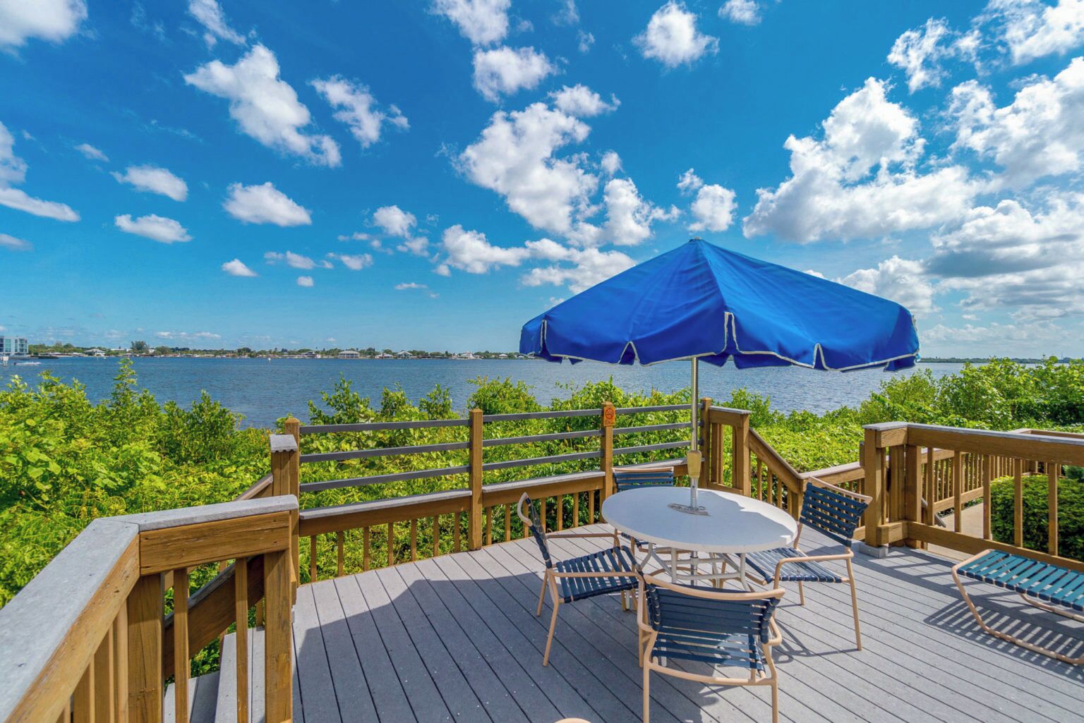 Um deck de madeira com mesa e cadeiras, um guarda-sol azul e vista para a água.