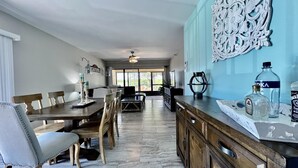 Un comedor amplio con una mesa y sillas de madera, una cocina con un detalle azul en la pared y vistas a través de puertas de cristal.
