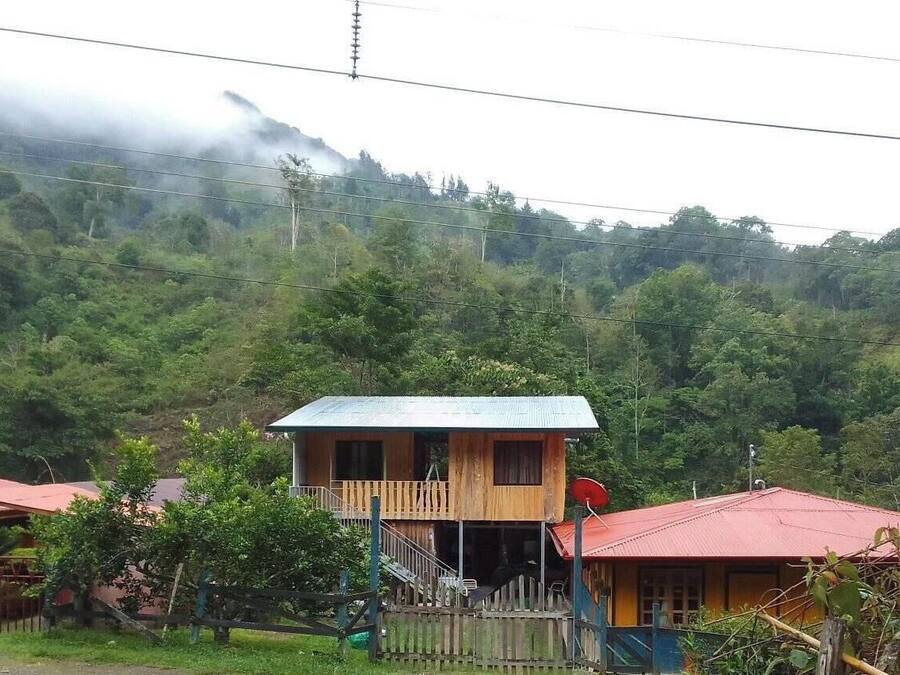 Una casa de madera con balcón, rodeada de abundante vegetación y montañas al fondo.