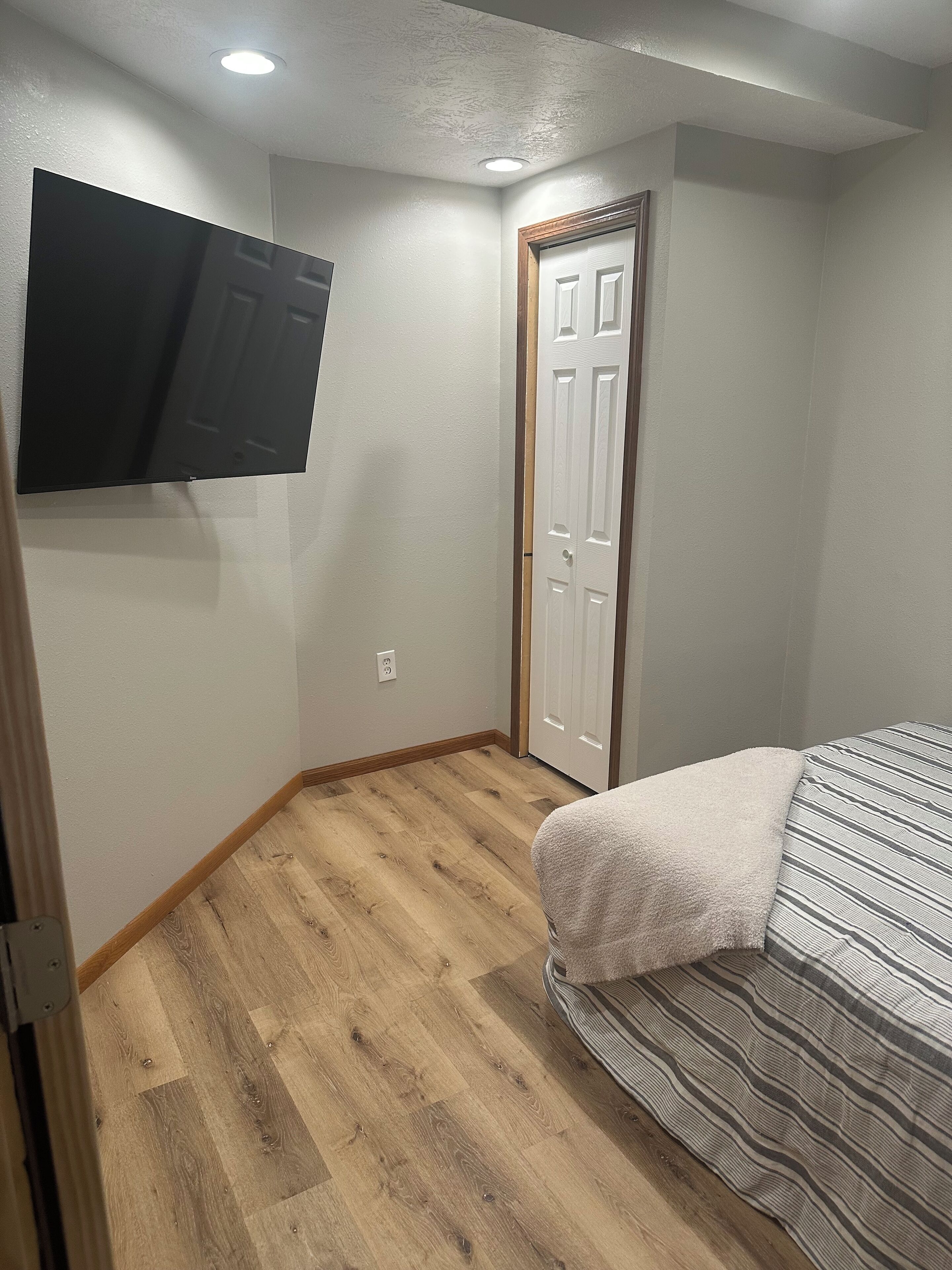 A bedroom with a bed, a flat-screen TV mounted on the wall, and a wooden floor.