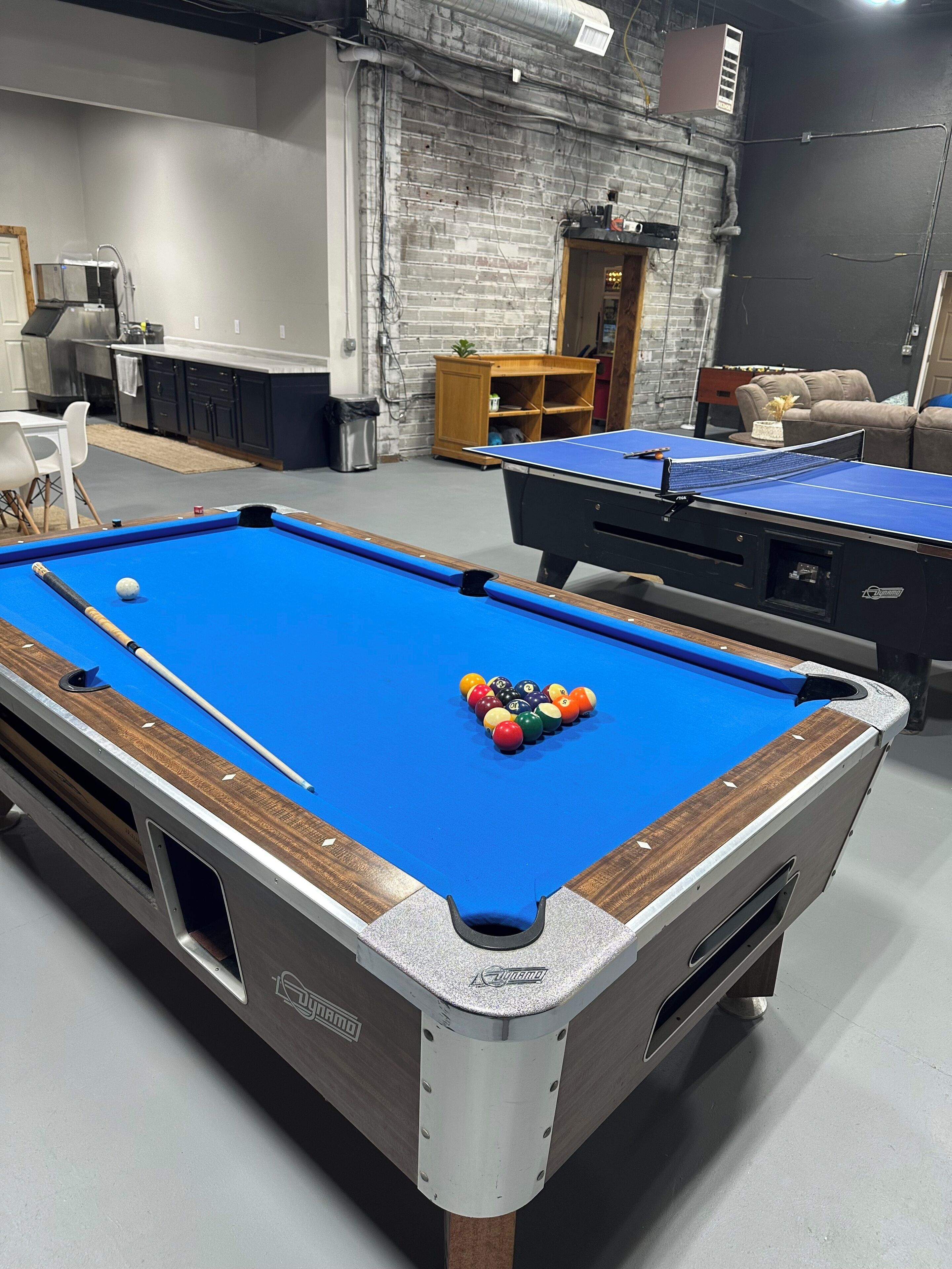 A pool table with balls and a cue stick, a second table in the background, and a brick wall with a door.