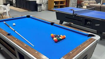 A pool table with balls and a cue stick, a second table in the background, and a brick wall with a door.
