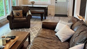 Un salón acogedor con un sofá de cuero marrón, una mesa de centro de madera y un sillón de cuero. Hay dos ventanas que dejan entrar luz natural y un ventilador de techo.