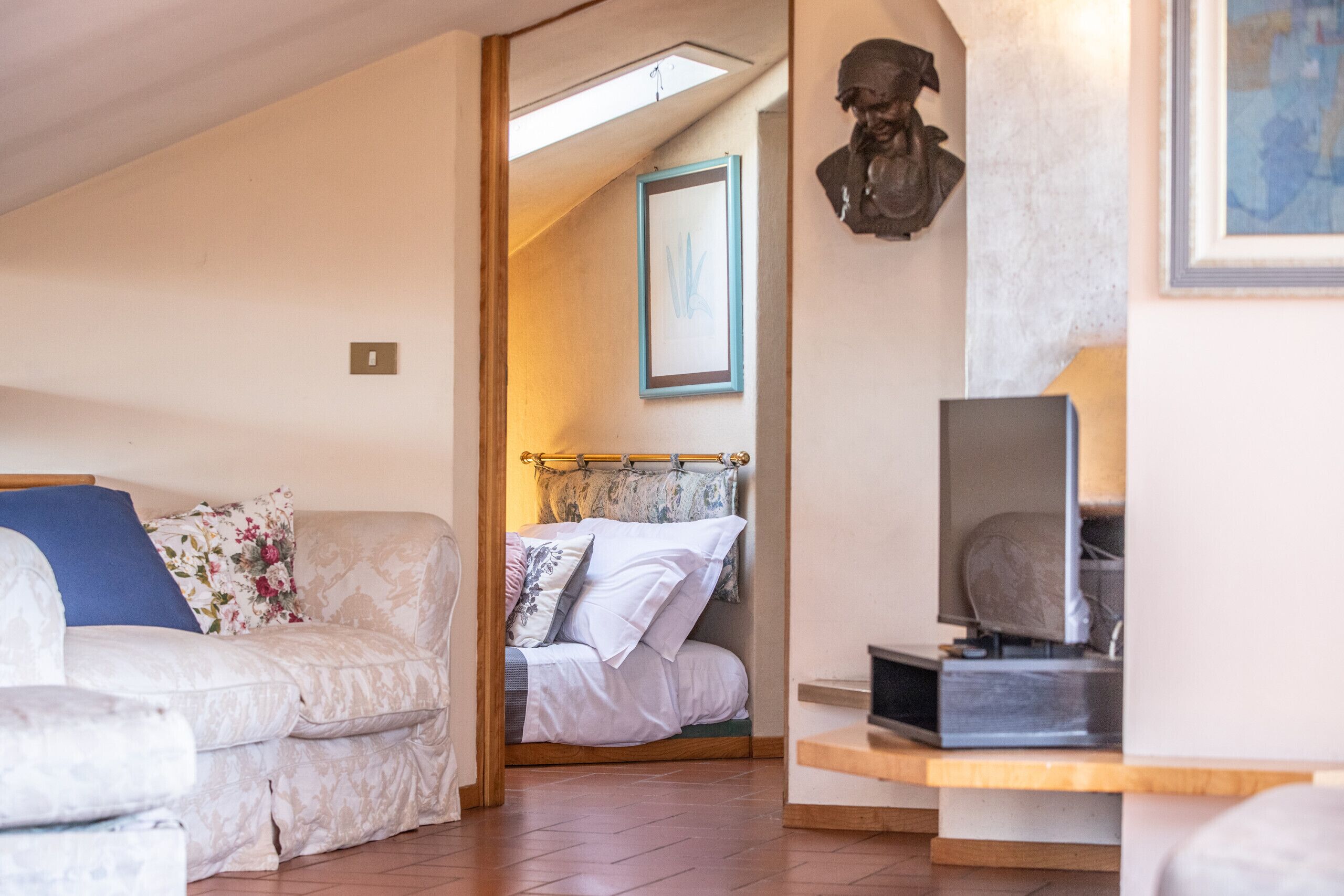 Una stanza accogliente con un divano, un letto con lenzuola a motivi floreali, una televisione e uno scultura a busto sulla parete.