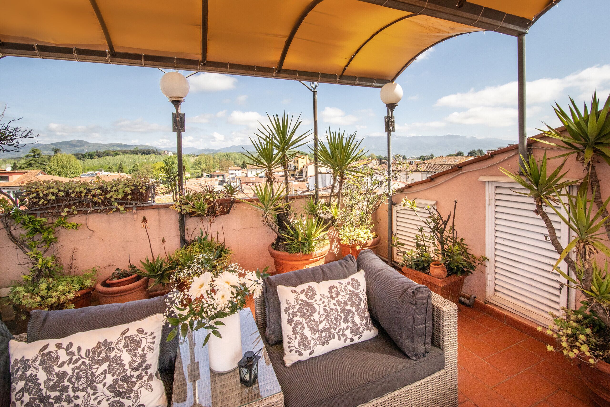 A covered outdoor seating area with potted plants, a wicker sofa, and a table with a lantern.