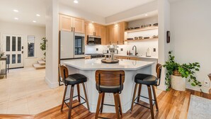 A modern kitchen with wooden cabinets, a central island with bar stools, and built-in appliances. - Broken Top Villa One - A Beautiful Two Bedroom Villa in Bend's Westside! (Bend)