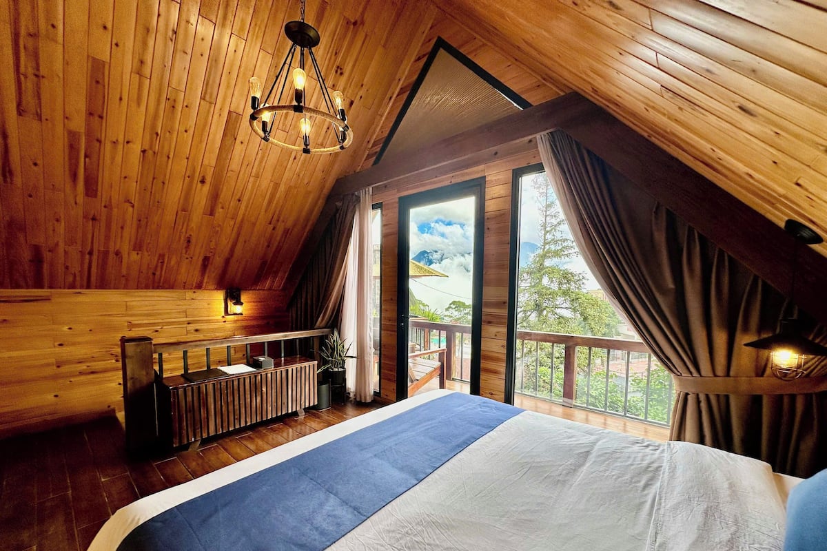 Ein Schlafzimmer in einer Holzhütte mit einem großen Bett, einer Holzbalkendecke und einem Balkon mit Blick auf Bäume.