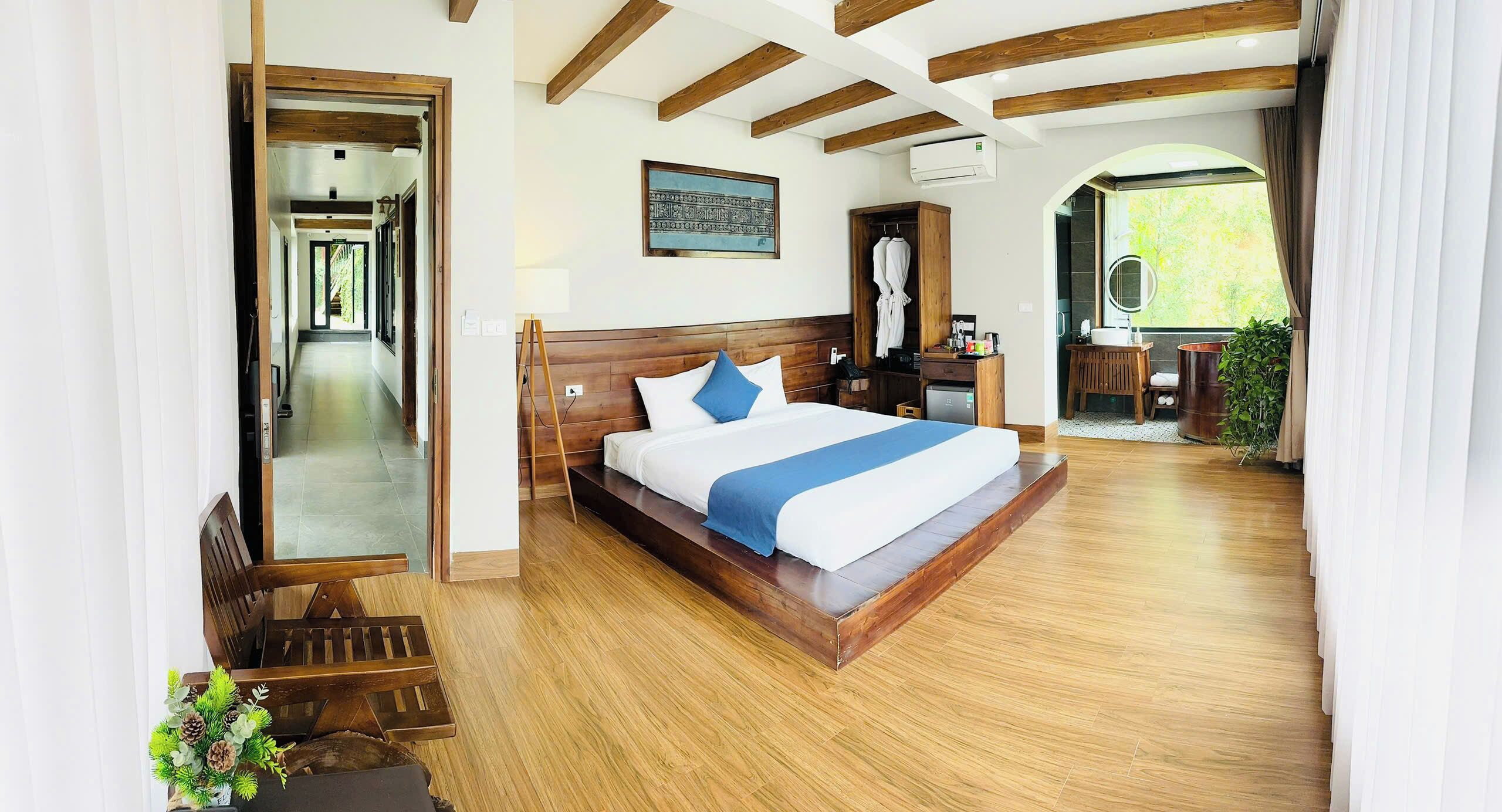 Un dormitorio amplio con una cama grande, vigas de madera, vistas a la vegetación y un baño con lavabo y espejo.
