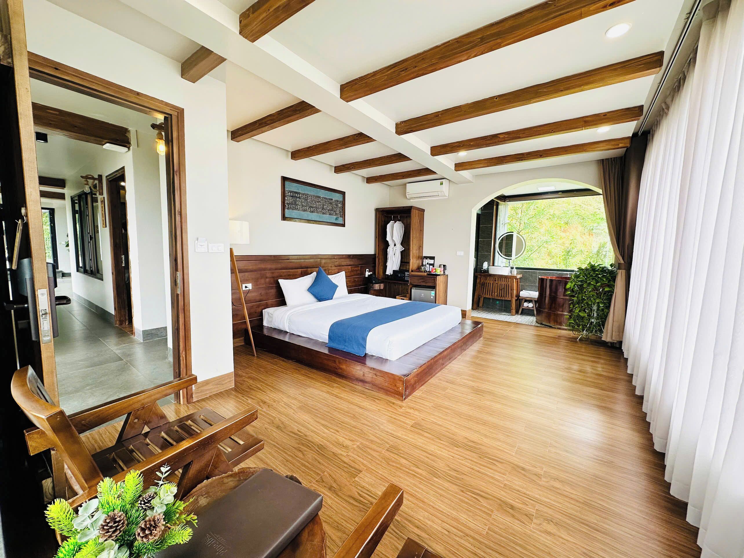 A spacious bedroom with a large bed, wooden beams on the ceiling, a wooden bench, a small desk, and a bathroom with a sink and mirror.
