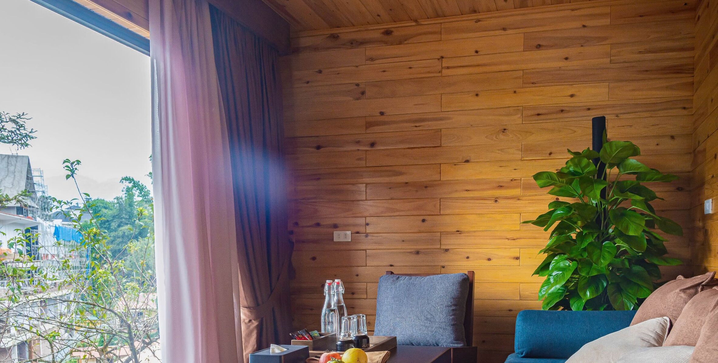 Een kamer met houten wanden, een groot raam met gordijnen, een blauwe bank, een grijze stoel en een groene plant.