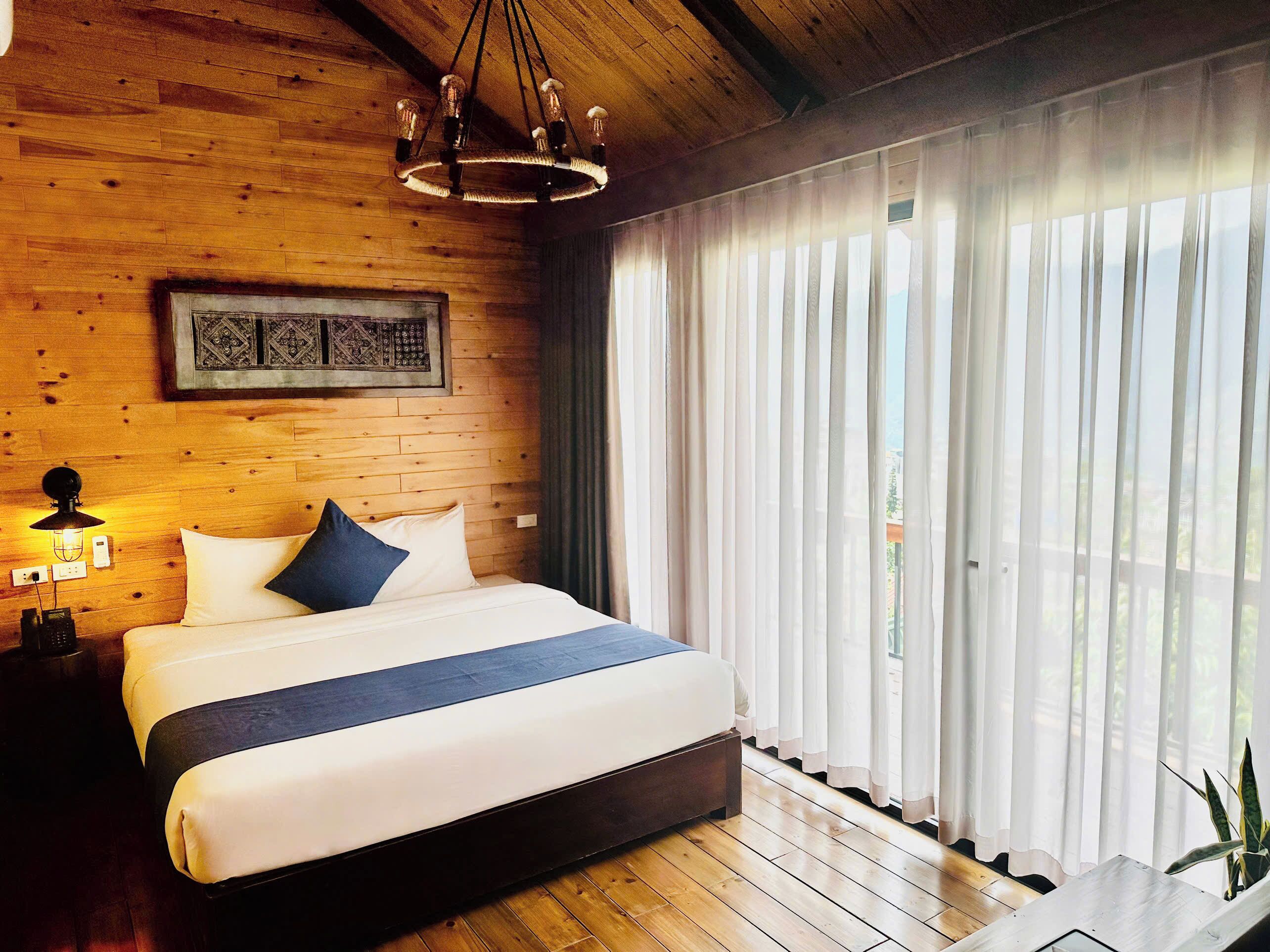 Une chambre à coucher aux murs lambrissés, avec un grand lit, un lustre, une lampe fixée au mur et une fenêtre ornée de rideaux légers.